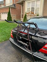 Side angled view showing the collapsible bike rack attached to a black sedan trunk with foam-padded arms and securing straps loose.