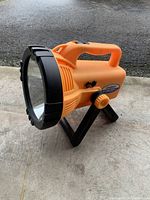 Side angle view of orange Sunforce portable light sitting on concrete surface showing handle, lens housing, and stand.