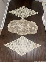 Three cream-colored hand embroidered Serbian table doilies arranged vertically on a dark wood floor, displaying detailed lace and crochet work, top doily diamond-shaped, middle oval with scene, bottom elongated with floral lace patterns.