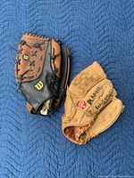 Two baseball mitts, one black and brown Wilson glove and one natural leather tan Rawlings glove with visible wear and writing on it.