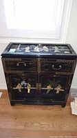 Front view of black lacquered cabinet showing two drawers and two doors with Asian themed artwork and metal hardware.