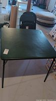 Square folding card table with green vinyl top and black metal folding legs shown in a basement setting.