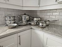 Kitchen corner countertop with stainless steel cookware arranged: two saucepans stacked, stock pot, medium pot, sauté pan, milk pan, two frying pans