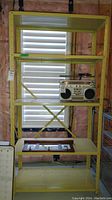 Full front view of yellow metal shelving unit showing five shelves, a stereo placed on the middle shelf and an open frame on the bottom shelf. Background has unfinished wall.