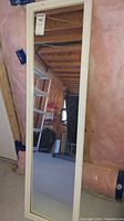 Full-length framed mirror standing vertically against basement wall with visible studs and insulation in the background, showing clear reflection of basement interior.