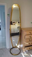 Front view of the tall oval-shaped mirror with brass frame standing next to a wooden dresser and reflecting a bedroom interior.