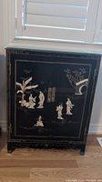 Front view of black lacquered wood cabinet showing Chinese painted motifs and stone figure accents on doors, metal pulls visible