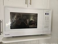 White Panasonic microwave oven placed on a kitchen shelf, front view showing digital clock display and control panel.