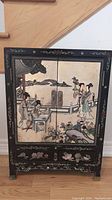 Front view of small black cabinet with two doors featuring painted scene of Asian women and flowers, lower drawers with floral designs, standing on floor.