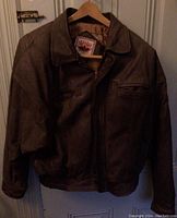 Front view of brown leather bomber jacket hanging on a wooden hanger.