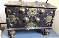 Front view showing dark brown wooden chest with brass dome studs and ornate brass plates on front panel and top