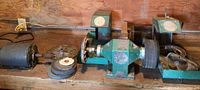 Wide shot showing two green bench grinders and an electric motor with various polishing wheels on a wooden surface against a plywood wall.