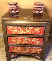 Front angled view of the vintage Asian cabinet showing the square top, three drawers with red and gold floral carved panels, brass pulls, and decorative feet. Two carved candle holders sit on top.