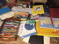 Photo showing an assortment of colored pencils, pens, various drawing pads by Blick and other brands, postcards and stencils scattered on a table.