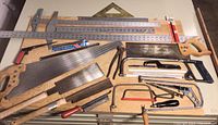 Photo showing a collection of hand saws with wooden handles, multiple metal rulers, steel squares, and small hand tools laid on a wooden surface.