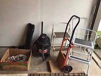 Full view of lot items including Shop Vac, hand truck, two step stools, and soft-sided box (partially open) with extension cords
