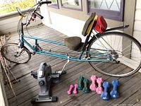 Full view of the vintage teal recumbent bike with padded seat and backrest, pedal exerciser attached, four sets of colored women's free weights on ground in front, and repair accessories.