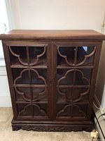Full view of the vintage wooden cabinet with glass front and carved patterns on doors and base.