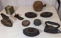 Wide photo of all items together on a cloth surface showing irons, teapot, stoneware box, tin, and match holder.