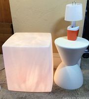 Front view of illuminated white plastic cube, white geometric small table, and battery lamp on top of table.
