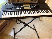 Yamaha electric keyboard on adjustable X-stand placed on tiled floor near window. Included headphones visible on keyboard.