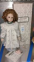 Porcelain doll with curly reddish-brown hair dressed in lace and embroidered white dress in front of box labeled Beautiful Dreamers.