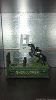 Front view of the cast iron mechanical bank showing the horse and rider in mid-jump on a green base labeled 'SHOWJUMPER'.