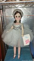 Front view of Elise Madame Alexander doll standing, dressed in silver sparkly dress and headpiece, holding tag.