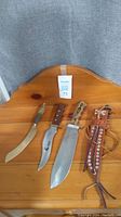 View of three knives laid out on wood surface with leather sheath alongside.