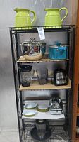 Metal shelving holding all kitchenware items described including two green ceramic pitchers on top shelf, pots and pan on second shelf, blenders on third shelf, dishes and utensils on lower shelves.