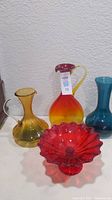 Wide shot showing all four items: yellow pitcher, amber to red tall pitcher with yellow handle, blue pitcher, and red glass bowl with wavy rim.
