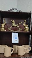Photo of two brass elephant statues and one brass rocking horse on a wooden shelf, with five beige coffee mugs below.