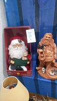 Photo of an animated Santa Claus figure in original box holding a green book and teddy bear, and a 16-inch clay Santa Claus statue holding a black bird and a small snowman.