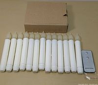 Photo of 13 white battery-operated LED candles with flame-shaped tops and a small brown box behind them, next to a silver remote control.