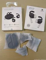 Two boxed sets of AirPods Pro ear hooks, each box labeled '3 Pairs Premium'. Loose black silicone ear hooks partially wrapped in translucent plastic foil in front of the boxes.