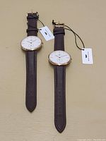 Two Brigada Swiss watches laid flat on a beige surface, showing brown leather straps and white dials with rose gold bezels