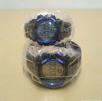 Two blue digital wristwatches wrapped in clear plastic, stacked on top of each other showing digital displays and blue bezels.