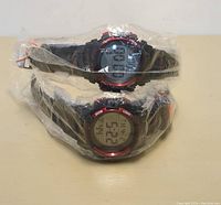 Two black digital watches with red accents, wrapped in protective plastic, showing digital displays on the faces.