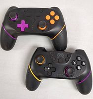 Front view of two wireless game controllers in black with colorful buttons and joystick accents, arranged side by side showing front button layouts and ergonomic grips.