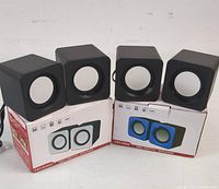 Two sets of mini digital speakers shown front and sides, with packaging boxes. Left: white and black speakers YST-1042. Right: blue and black speakers YST-1052.
