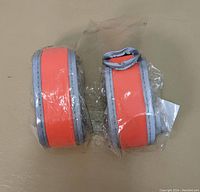 Two orange wristband reflectors with reflective silver edges, individually wrapped in plastic, side by side on a beige surface.
