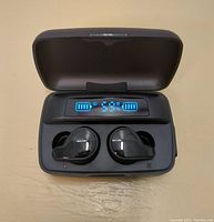 Black wireless Bluetooth earbuds placed inside the charging case, digital LED screen showing battery levels of left and right earbuds.