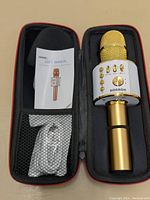 Gold and white wireless Bluetooth karaoke microphone and accessories inside a black and red zippered carrying case. The case contains the microphone, a user manual, and a white charging cable in plastic packaging.