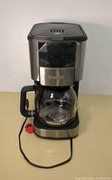 Front view of Brewsly coffee maker showing stainless steel and black plastic body with glass carafe and power cord.
