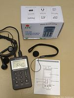 Digital handheld HRD FM/AM stereo DSP receiver powered on showing 640 frequency, wired earphones plugged in, wrist strap attached, instruction manual and product box in background.