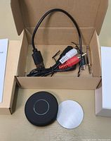 Photo of the SONRU Mini Wireless 5.0 Music Receiver and included accessories inside an open cardboard box, showing the receiver, RCA cables, USB charging cable, and adhesive pad.
