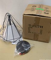 Conical geometric wireframe pendant light next to a cardboard box and black ceiling mount in plastic