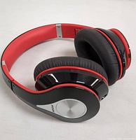 Close-up side angle view of red and black Mpow wireless over-ear headphones showing cushioned ear pads, silver circular design on ear cup, and padded red headband.