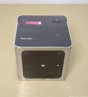 Front and top view of Petcube smart pet camera inside clear protective wrapping, showing camera lens and Petcube logo.