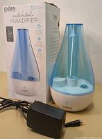 Photo showing the humidifier unit with transparent blue water tank, white base, power button, and included black power adapter cable.